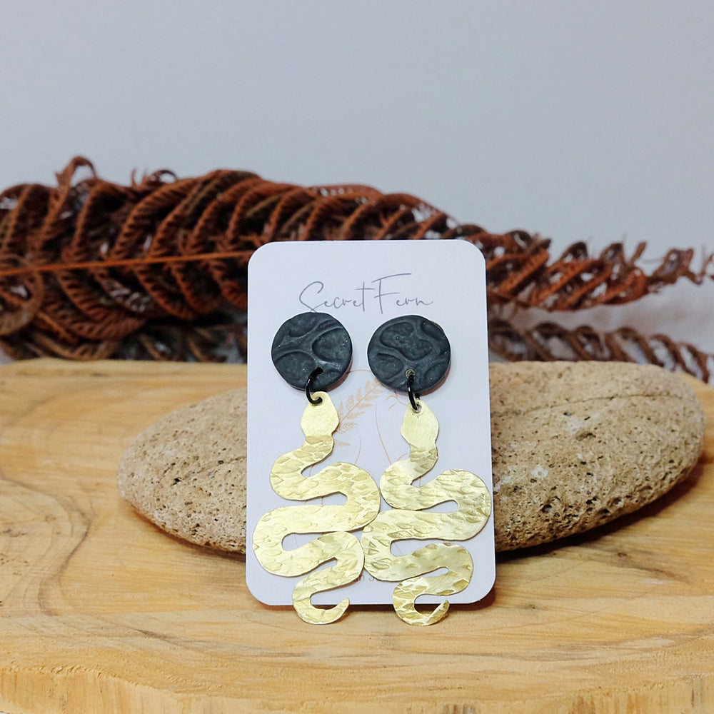 
            
                Load image into Gallery viewer, textured brass snake earrings on a black textured stud
            
        