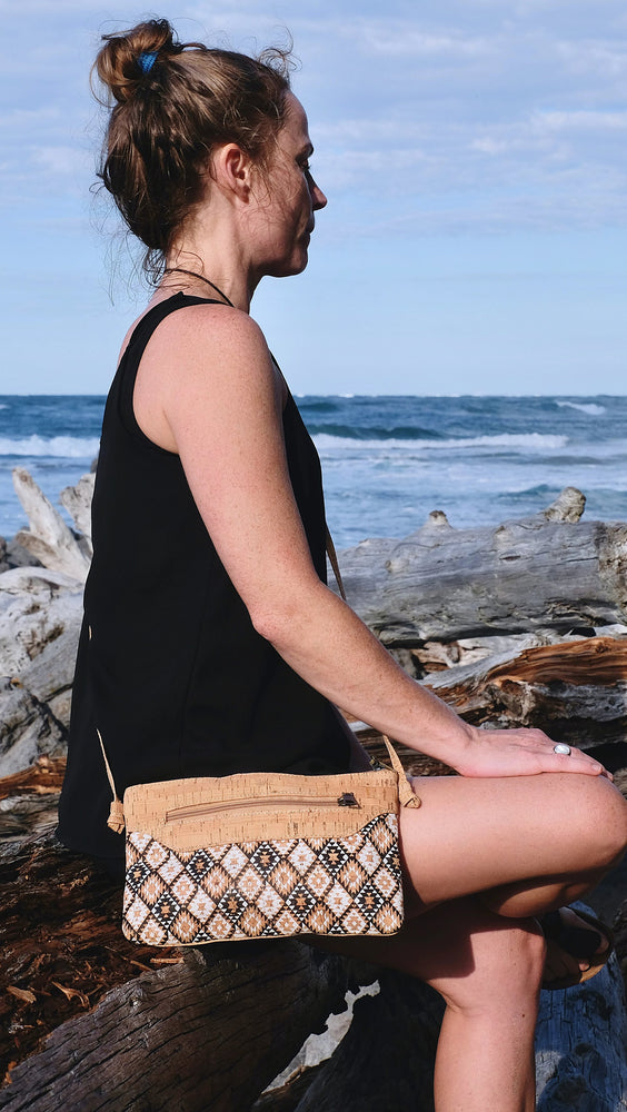 Model posing at the beach wearing a triangular boho patterns and cork vegan fabric, this minimalist pouch bag. Made with eco-friendly, sustainable, PETA approved and fair-trade European standards. 
